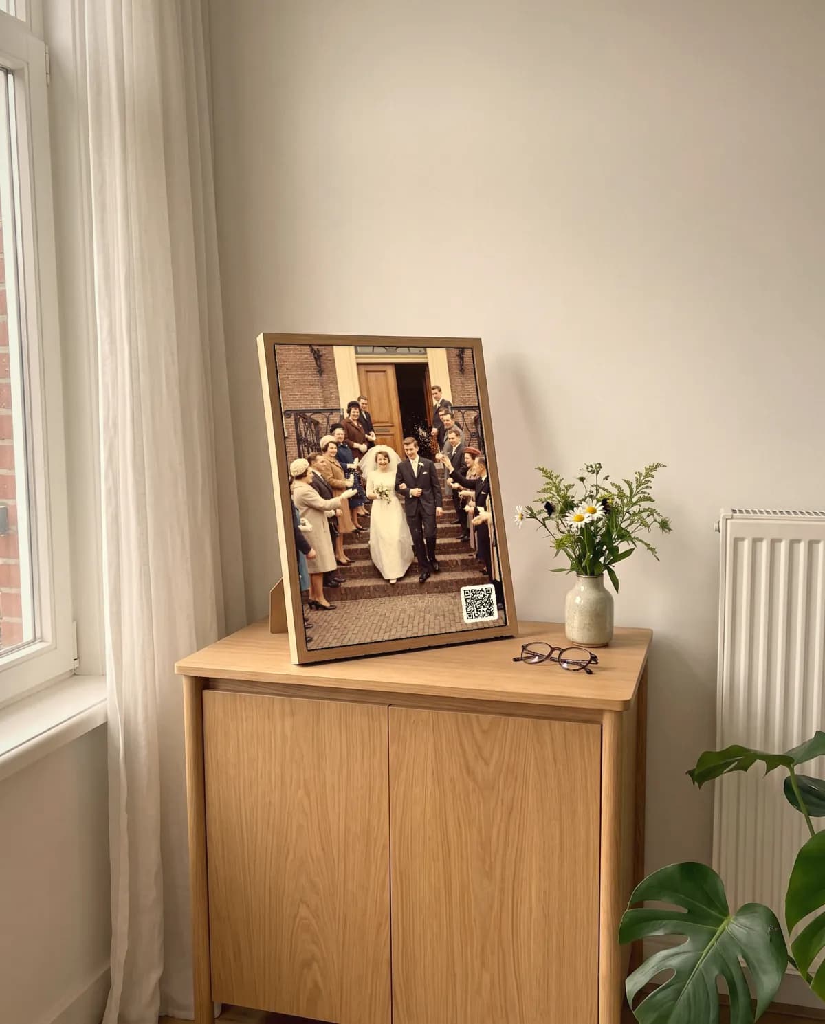 Un Cadre à histoires avec une photo de mariage de 1962 et un QR code dans le coin, posé sur un petit meuble en chêne dans un salon lumineux